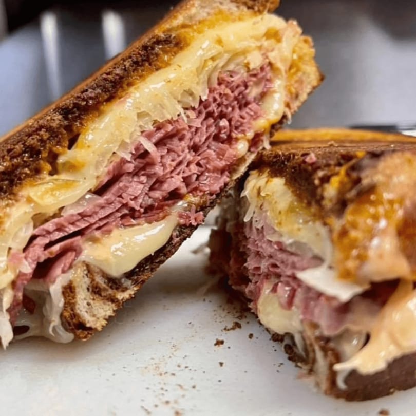 Close-up of a grilled sandwich, cut in half, revealing layers of pastrami, melted cheese, and sauerkraut.