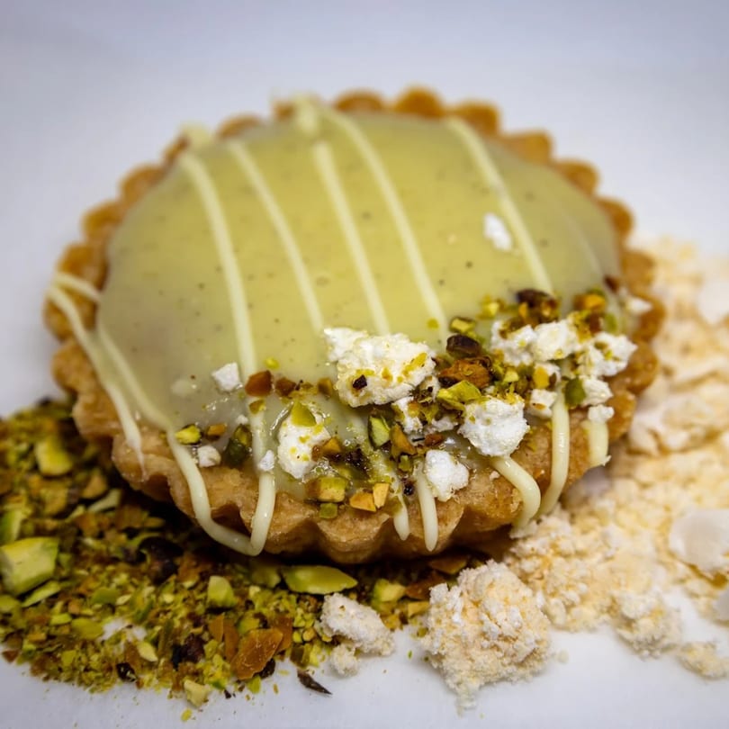 A pistachio tart with white chocolate drizzle and crumbled pistachios on a white surface.