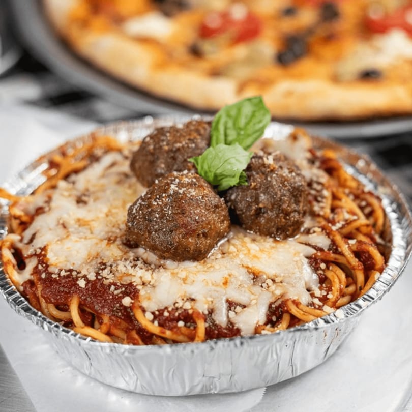 Spaghetti and meatballs with melted cheese and basil in a foil container, with a pizza in the background.